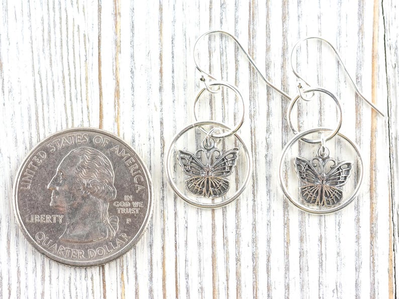 May include: Silver butterfly earrings with a quarter coin for size comparison. The earrings feature a detailed butterfly design within a circular frame, suspended from a hook. The background is a white wooden surface.