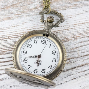 May include: A vintage-style pocket watch with a gold-toned metal case and a white face. The watch has black Roman numeral hour markers and black hands. The watch is open and the chain is visible.