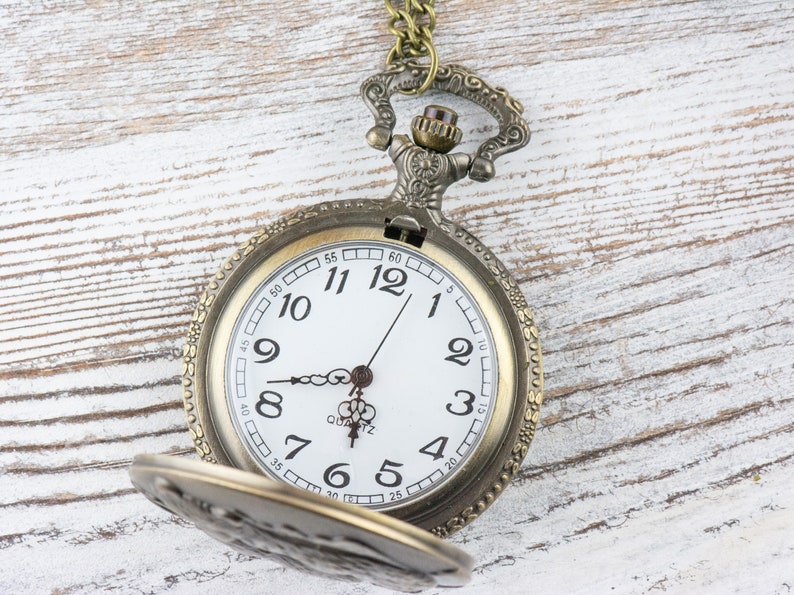 May include: A vintage-style pocket watch with a gold-toned metal case and a white face. The watch has black Roman numeral hour markers and black hands. The watch is open and the chain is visible.