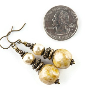 K&ouml;nnte beinhalten: Ein Paar goldfarbene Ohrringe mit einer gro&szlig;en, unregelm&auml;&szlig;ig geformten, cremefarbenen Perle und einer kleineren cremefarbenen Perle. Die Ohrringe werden neben einer 20-Cent-M&uuml;nze zum Gr&ouml;&szlig;envergleich gezeigt.