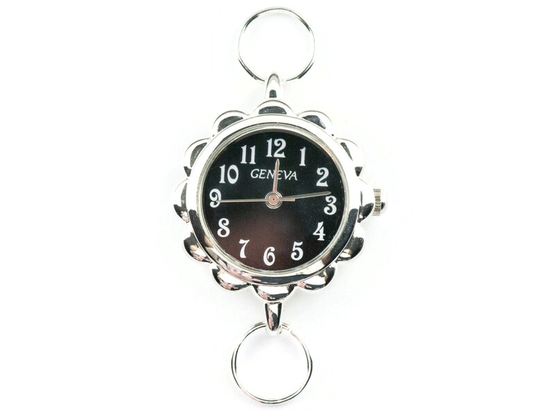 May include: A silver-toned watch with a black face and white numbers. The watch has a decorative, scalloped edge and is attached to a double ring.
