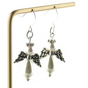 May include: Silver angel wing earrings with a white pearl bead dangle. The wings are detailed with black accents.
