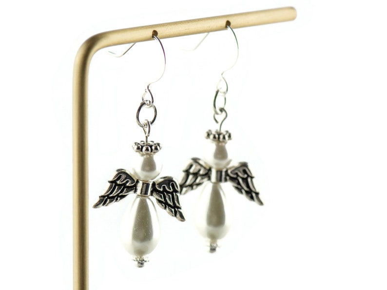 May include: Silver angel wing earrings with a white pearl bead dangle. The wings are detailed with black accents.