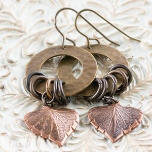 Op de afbeelding: Een paar handgemaakte oorbellen met grote, gehamerde messing hoepels en kleinere, verbonden metalen ringen in brons- en zilverkleuren. Elke oorbel heeft een koperen blad-vormige bedel met gedetailleerde adering.
