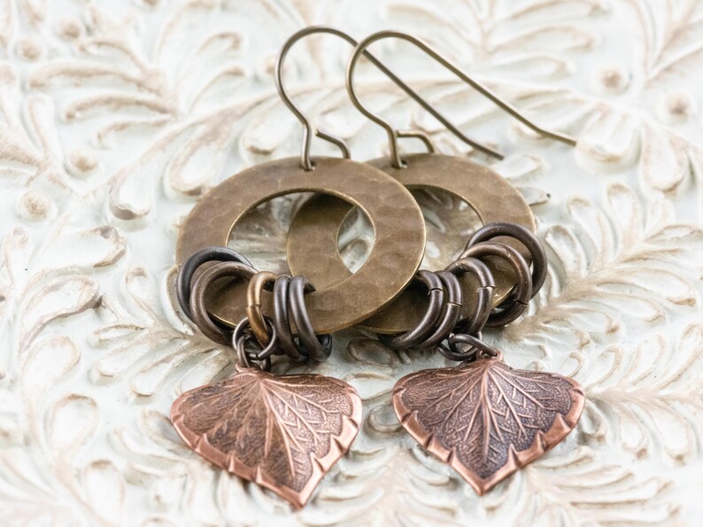 Op de afbeelding: Een paar handgemaakte oorbellen met grote, gehamerde messing hoepels en kleinere, verbonden metalen ringen in brons- en zilverkleuren. Elke oorbel heeft een koperen blad-vormige bedel met gedetailleerde adering.