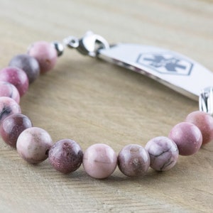 May include: A pink and gray beaded bracelet with a silver medical ID tag. The tag has a medical symbol and is engraved with text.