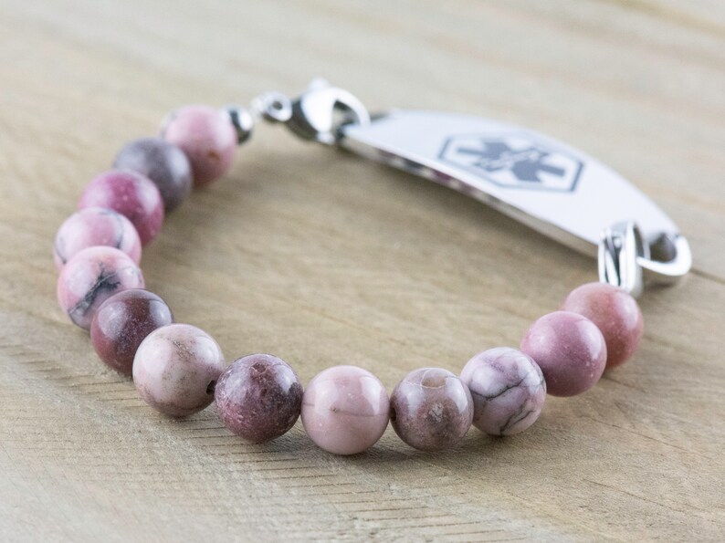 May include: A pink and gray beaded bracelet with a silver medical ID tag. The tag has a medical symbol and is engraved with text.