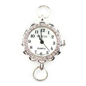 May include: A silver-toned Geneva quartz watch with a white face and black numerals. The watch has a floral-shaped bezel encrusted with clear crystals. It includes two silver rings for attachment.