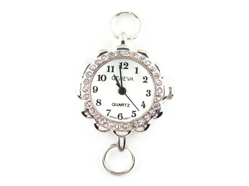 May include: A silver-toned Geneva quartz watch with a white face and black numerals. The watch has a floral-shaped bezel encrusted with clear crystals. It includes two silver rings for attachment.