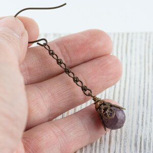 May include: A single antique bronze earring with a chain and a purple gemstone. The earring is held in a hand against a white wooden background. The gemstone is encased in a decorative bronze cap.