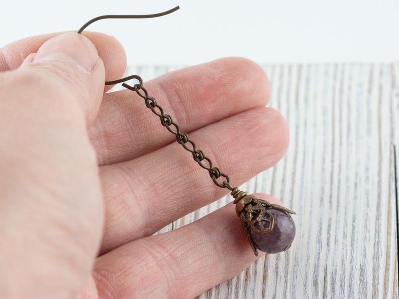 May include: A single antique bronze earring with a chain and a purple gemstone. The earring is held in a hand against a white wooden background. The gemstone is encased in a decorative bronze cap.