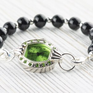 Op de afbeelding: Een groene wijzerplaat met zwarte Romeinse cijfers en een zilveren horlogekast. Het horloge is bevestigd aan een zwarte kralenarmband.