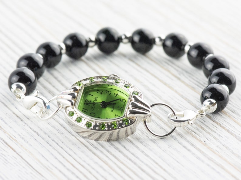 Op de afbeelding: Een groene wijzerplaat met zwarte Romeinse cijfers en een zilveren horlogekast. Het horloge is bevestigd aan een zwarte kralenarmband.