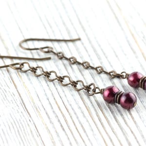 Puede incluir: Un par de pendientes colgantes con cuentas redondas de color rojo oscuro suspendidas de una cadena de peque&ntilde;os eslabones met&aacute;licos de color bronce. Los pendientes tienen un dise&ntilde;o de gancho simple.