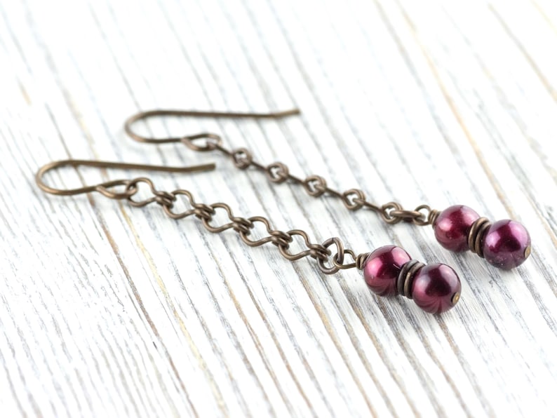 Puede incluir: Un par de pendientes colgantes con cuentas redondas de color rojo oscuro suspendidas de una cadena de peque&ntilde;os eslabones met&aacute;licos de color bronce. Los pendientes tienen un dise&ntilde;o de gancho simple.