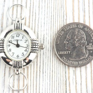 Pu&ograve; includere: Orologio Geneva argentato con quadrante bianco e numeri neri. L'orologio ha un quadrante rotondo e due anelli per il fissaggio. Una moneta &egrave; accanto all'orologio per il confronto delle dimensioni.