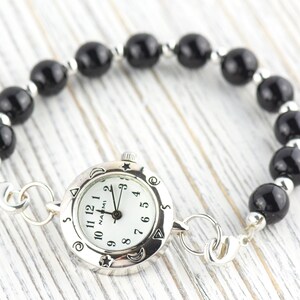 May include: A wristwatch with a round silver-tone case and a white dial featuring the text NAOMI, black numerals, and celestial symbols on the bezel. The watch is attached to a bracelet made of polished black spherical beads and small silver-tone spacers.