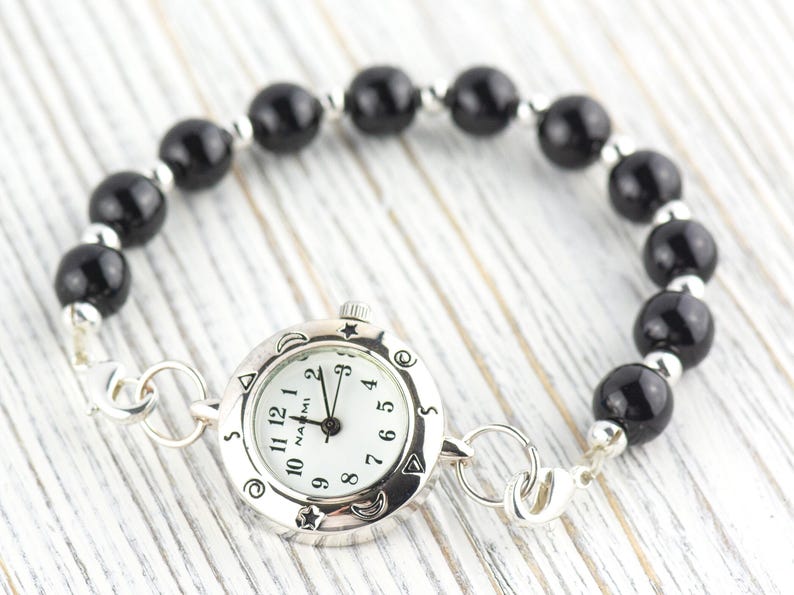 May include: A wristwatch with a round silver-tone case and a white dial featuring the text NAOMI, black numerals, and celestial symbols on the bezel. The watch is attached to a bracelet made of polished black spherical beads and small silver-tone spacers.
