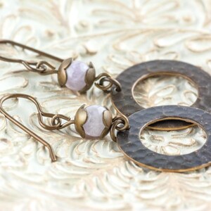 Puede incluir: Pendientes hechos a mano con una cuenta de piedra preciosa de color p&uacute;rpura claro y un anillo de metal color bronce martillado. Los pendientes tienen una est&eacute;tica vintage y se exhiben sobre una superficie texturizada de color blanquecino.