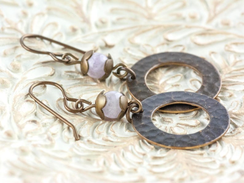 Puede incluir: Pendientes hechos a mano con una cuenta de piedra preciosa de color p&uacute;rpura claro y un anillo de metal color bronce martillado. Los pendientes tienen una est&eacute;tica vintage y se exhiben sobre una superficie texturizada de color blanquecino.