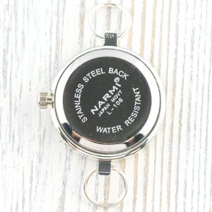 K&ouml;nnte beinhalten: Eine silberfarbene Taschenuhr mit schwarzem Zifferblatt. Auf der R&uuml;ckseite der Uhr steht "STAINLESS STEEL BACK" und "NARMI JAPAN MOVT L-106 WATER RESISTANT". Die Uhr hat zwei Metallschlaufen und eine Aufzugswelle.