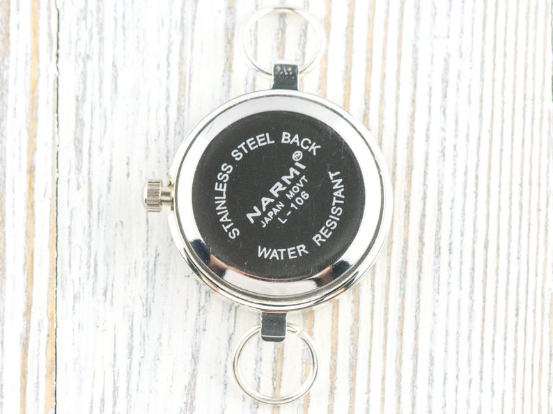 K&ouml;nnte beinhalten: Eine silberfarbene Taschenuhr mit schwarzem Zifferblatt. Auf der R&uuml;ckseite der Uhr steht "STAINLESS STEEL BACK" und "NARMI JAPAN MOVT L-106 WATER RESISTANT". Die Uhr hat zwei Metallschlaufen und eine Aufzugswelle.