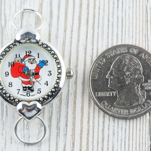 Peut inclure: Une montre argent&eacute;e avec un cadran blanc repr&eacute;sentant un P&egrave;re No&euml;l dessin anim&eacute; tenant un sac de cadeaux. La montre a une cha&icirc;ne argent&eacute;e et un fermoir en forme de c&oelig;ur. La montre est &agrave; c&ocirc;t&eacute; d'une pi&egrave;ce de monnaie am&eacute;ricaine de 25 cents.