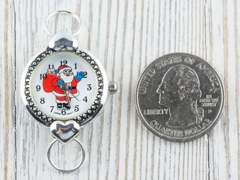 Peut inclure: Une montre argent&eacute;e avec un cadran blanc repr&eacute;sentant un P&egrave;re No&euml;l dessin anim&eacute; tenant un sac de cadeaux. La montre a une cha&icirc;ne argent&eacute;e et un fermoir en forme de c&oelig;ur. La montre est &agrave; c&ocirc;t&eacute; d'une pi&egrave;ce de monnaie am&eacute;ricaine de 25 cents.