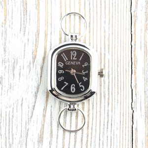 May include: A silver-toned Geneva pocket watch with a black face and white numerals. The watch has a rectangular shape with rounded corners and two circular rings for attachment. The watch is displayed on a white wooden surface.