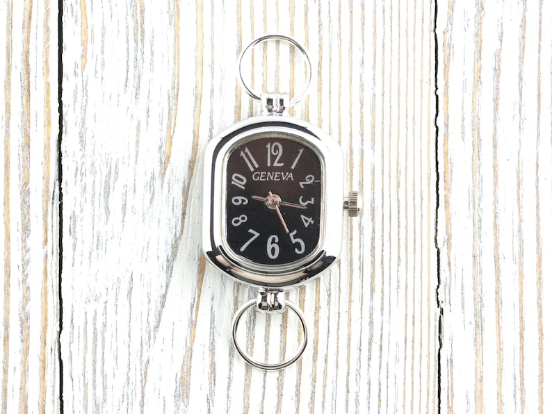 May include: A silver-toned Geneva pocket watch with a black face and white numerals. The watch has a rectangular shape with rounded corners and two circular rings for attachment. The watch is displayed on a white wooden surface.