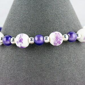 May include: A purple and white beaded bracelet with floral designs on the white beads.