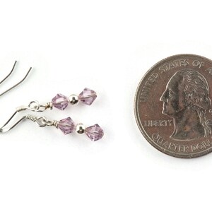 May include: A pair of delicate silver earrings with lavender-colored crystal beads. The earrings are displayed next to a U.S. quarter for size comparison. The crystals are faceted, reflecting light. The earrings have a simple, elegant design.