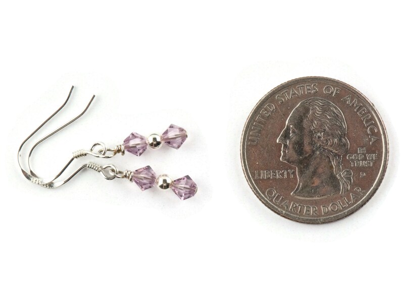 May include: A pair of delicate silver earrings with lavender-colored crystal beads. The earrings are displayed next to a U.S. quarter for size comparison. The crystals are faceted, reflecting light. The earrings have a simple, elegant design.