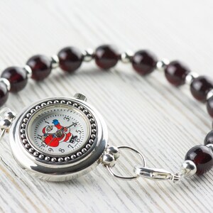 Puede incluir: Un reloj plateado con una esfera blanca y un dise&ntilde;o de Pap&aacute; Noel. El reloj est&aacute; unido a una pulsera hecha de cuentas de color rojo oscuro.