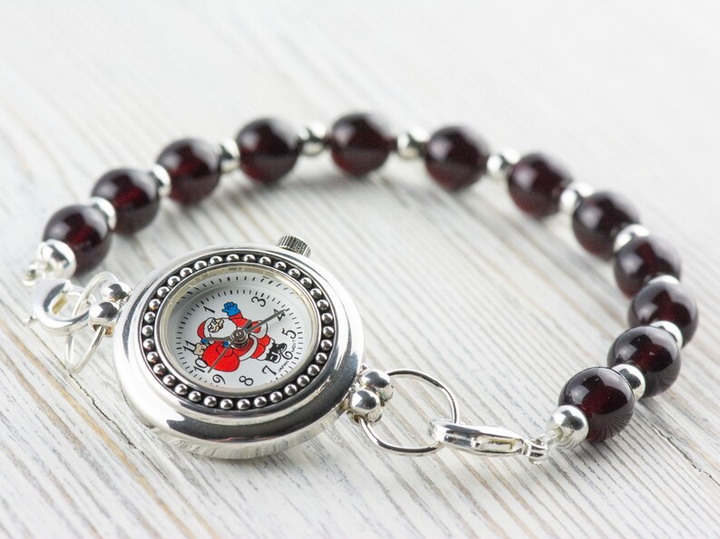 Puede incluir: Un reloj plateado con una esfera blanca y un dise&ntilde;o de Pap&aacute; Noel. El reloj est&aacute; unido a una pulsera hecha de cuentas de color rojo oscuro.