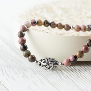 Puede incluir: Un brazalete de cuentas de color rosa y marr&oacute;n con un dije de plata con un dise&ntilde;o decorativo. El brazalete est&aacute; sobre una superficie blanca.