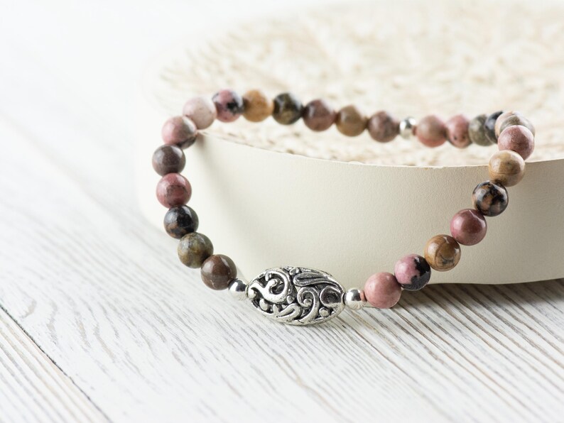 Puede incluir: Un brazalete de cuentas de color rosa y marr&oacute;n con un dije de plata con un dise&ntilde;o decorativo. El brazalete est&aacute; sobre una superficie blanca.
