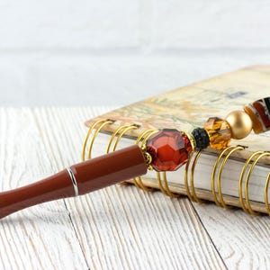 Puede incluir: Un bol&iacute;grafo marr&oacute;n con punta plateada y una parte superior decorativa con cuentas. Las cuentas incluyen una gran gema roja facetada, detalles dorados, &aacute;mbar y negros. El bol&iacute;grafo descansa sobre un cuaderno con espiral con anillos dorados.