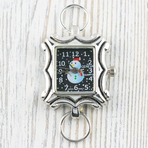 May include: A silver-toned pocket watch with a black face and a snowman design. The watch has a key ring attached to it and reads "Geneva Quartz" and "Japan Movt".