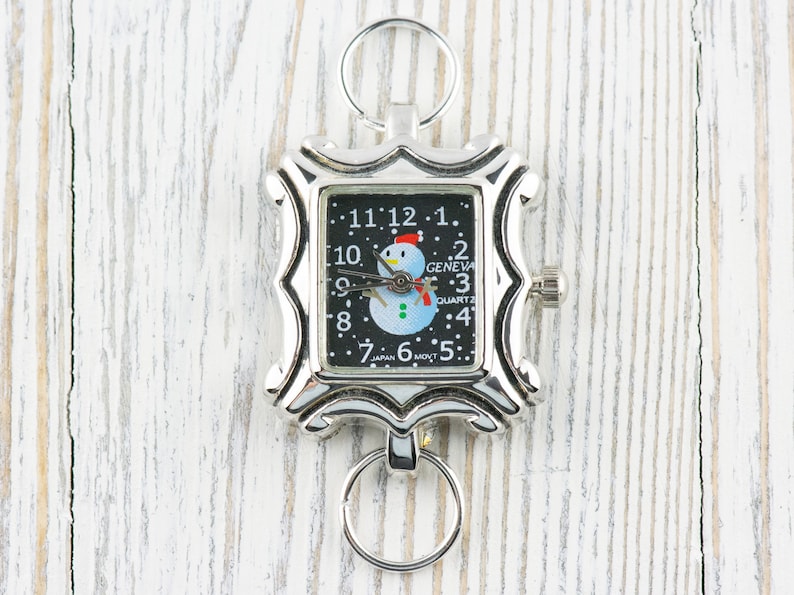 May include: A silver-toned pocket watch with a black face and a snowman design. The watch has a key ring attached to it and reads "Geneva Quartz" and "Japan Movt".