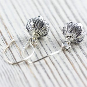 Pu&ograve; includere: Un paio di orecchini d'argento con un design floreale. Ogni orecchino presenta una perla d'argento a forma di fiore, dettagliata e strutturata, sospesa a un semplice gancio d'argento. Gli orecchini sono esposti su una superficie di legno di colore chiaro.