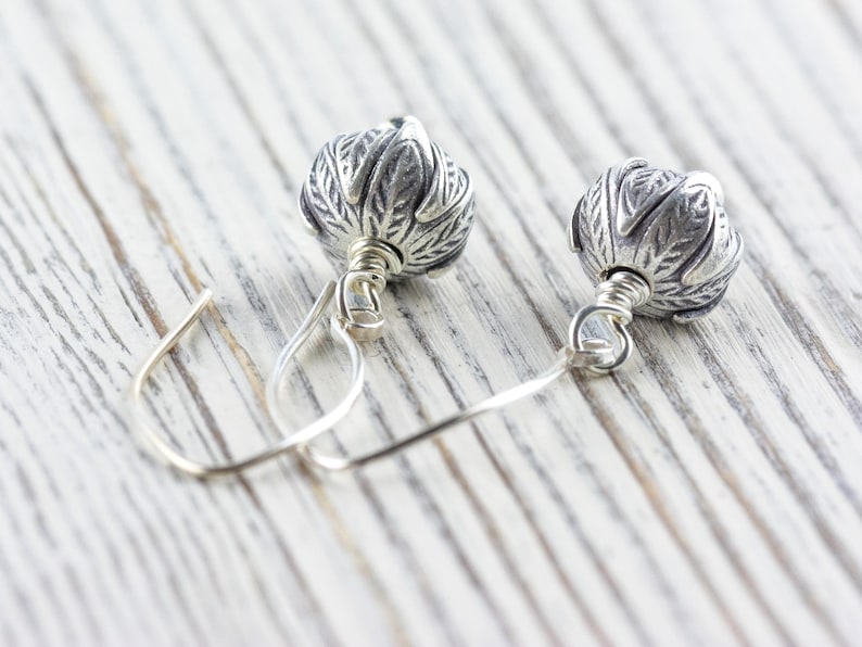 Pu&ograve; includere: Un paio di orecchini d'argento con un design floreale. Ogni orecchino presenta una perla d'argento a forma di fiore, dettagliata e strutturata, sospesa a un semplice gancio d'argento. Gli orecchini sono esposti su una superficie di legno di colore chiaro.