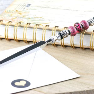 May include: A decorative letter opener with a silver blade and a beaded handle. The handle features pink leopard print beads, black beads, and silver accents. A white envelope with a heart sticker and a spiral-bound notebook are in the background.