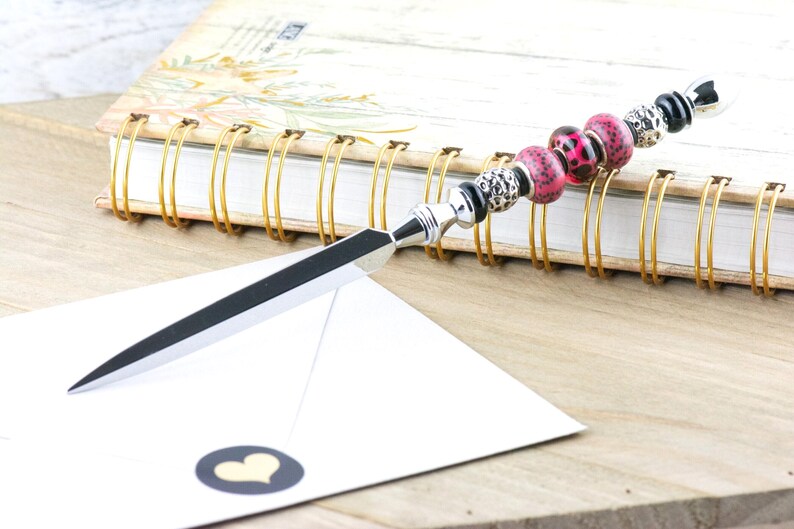 May include: A decorative letter opener with a silver blade and a beaded handle. The handle features pink leopard print beads, black beads, and silver accents. A white envelope with a heart sticker and a spiral-bound notebook are in the background.