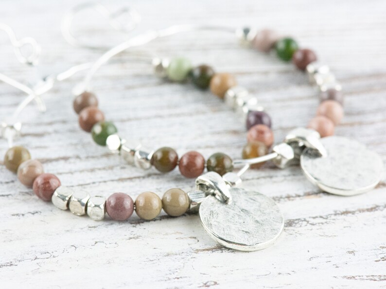 May include: A pair of silver hoop earrings with a silver charm pendant. The earrings are made with a variety of colorful stones, including green, brown, and pink.