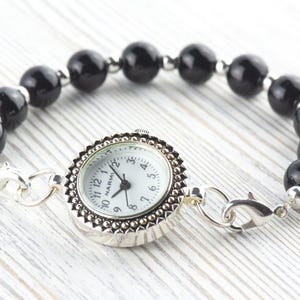 K&ouml;nnte beinhalten: Ein silberfarbenes Armband mit einem wei&szlig;en Zifferblatt und einem schwarzen Perlenband. Das Zifferblatt hat eine dekorative silberne L&uuml;nette und ein wei&szlig;es Zifferblatt mit schwarzen Ziffern. Das Armband ist mit silbernen Verschl&uuml;ssen verbunden.
