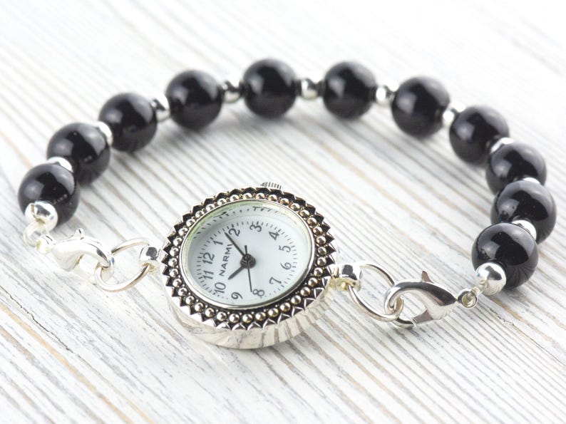 K&ouml;nnte beinhalten: Ein silberfarbenes Armband mit einem wei&szlig;en Zifferblatt und einem schwarzen Perlenband. Das Zifferblatt hat eine dekorative silberne L&uuml;nette und ein wei&szlig;es Zifferblatt mit schwarzen Ziffern. Das Armband ist mit silbernen Verschl&uuml;ssen verbunden.