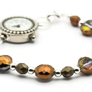 K&ouml;nnte beinhalten: Eine silberne Uhr mit wei&szlig;em Zifferblatt und schwarzen Zeigern, die an einem Armband mit braunen, goldenen und silbernen Perlen befestigt ist.