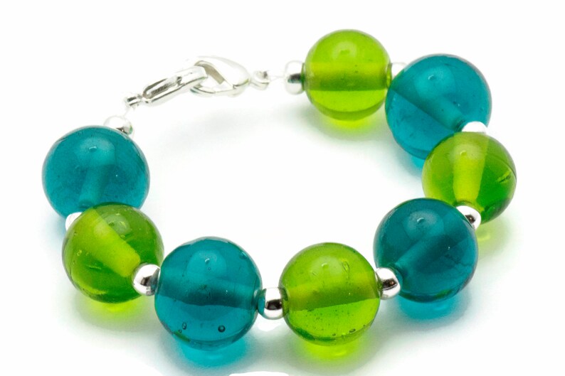 Puede incluir: Un brazalete hecho de cuentas de vidrio alternas de color verde azulado y verde lima con separadores plateados.