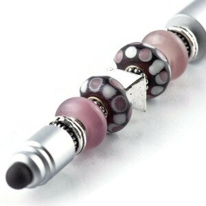 May include: A silver stylus pen with a black tip and a series of pink and purple glass beads with white polka dots.
